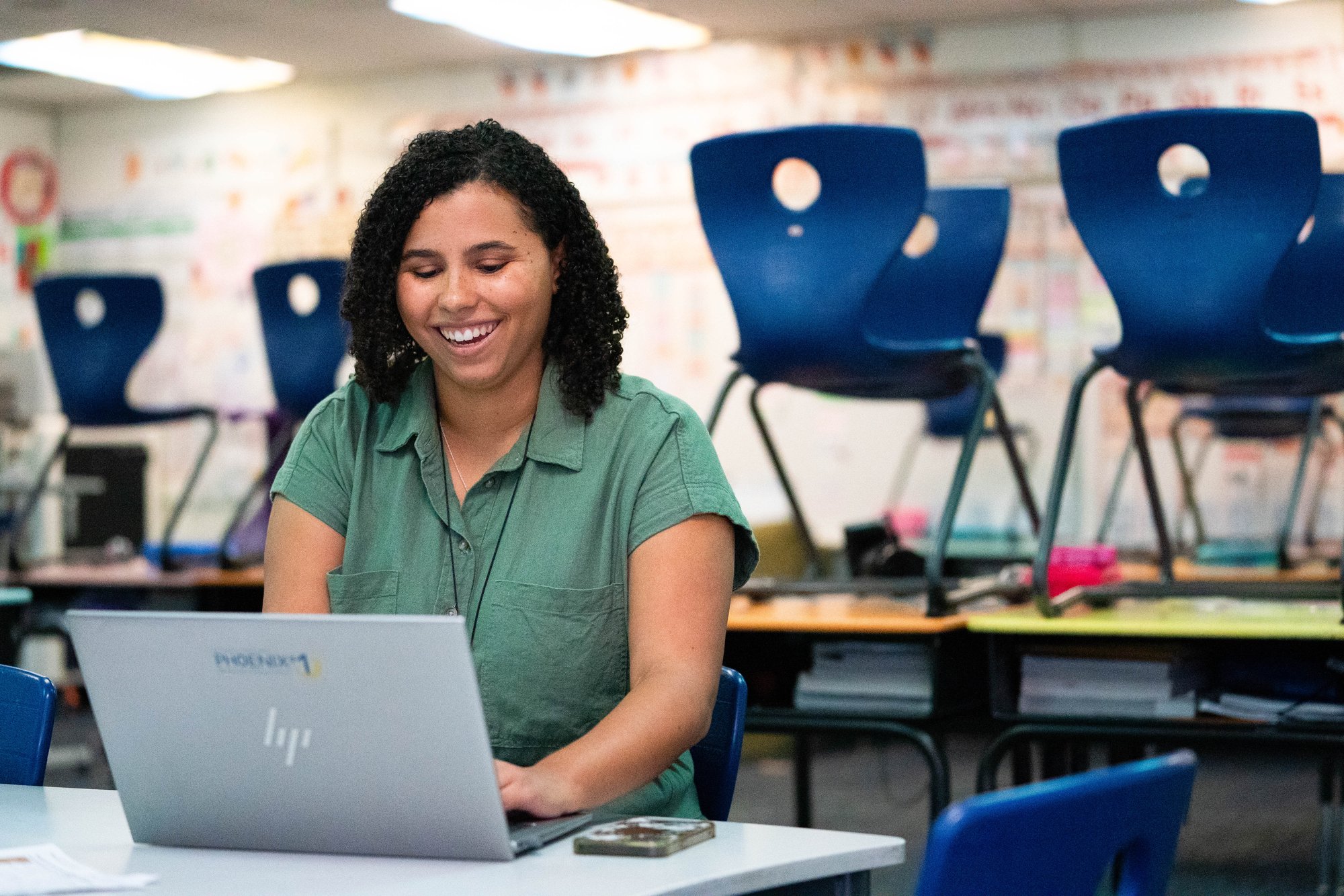 A smiling teacher sits at a desk typing on a laptop.