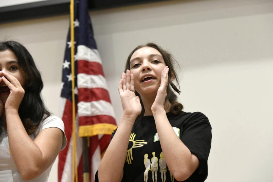 Students stand on a stage in front of an American flag shouting with enthusiasm