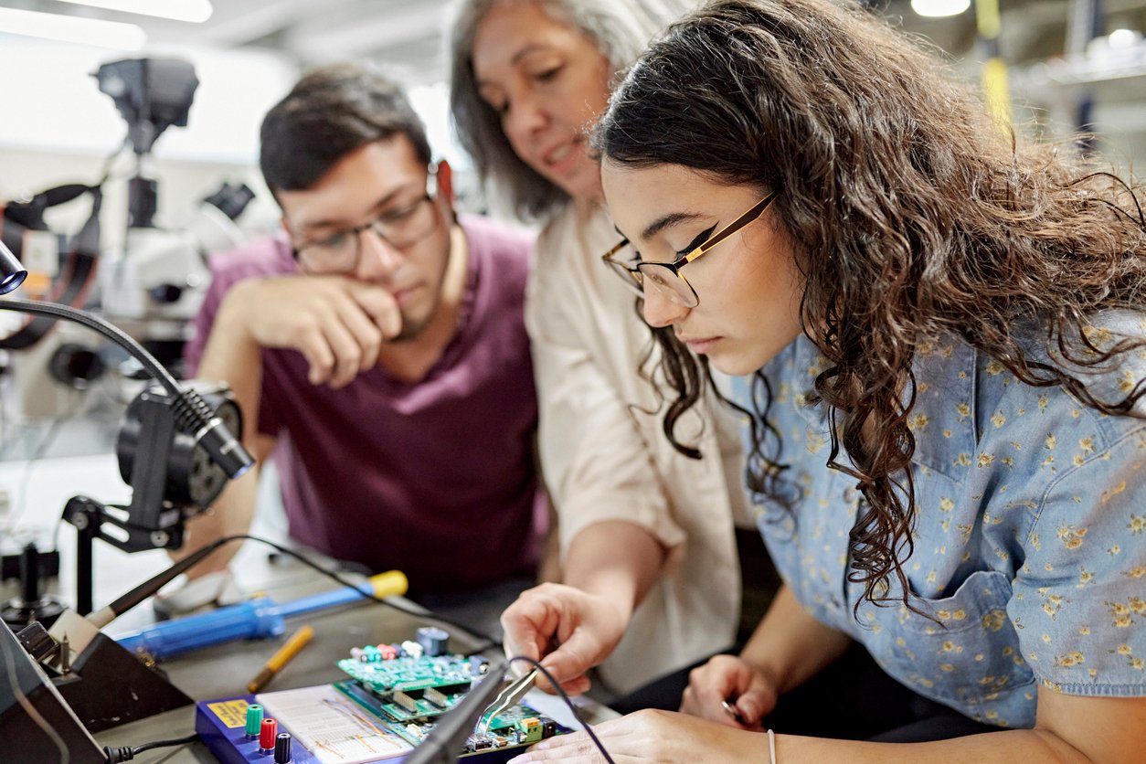 Two high school students and their teacher work intently using electronic equipment