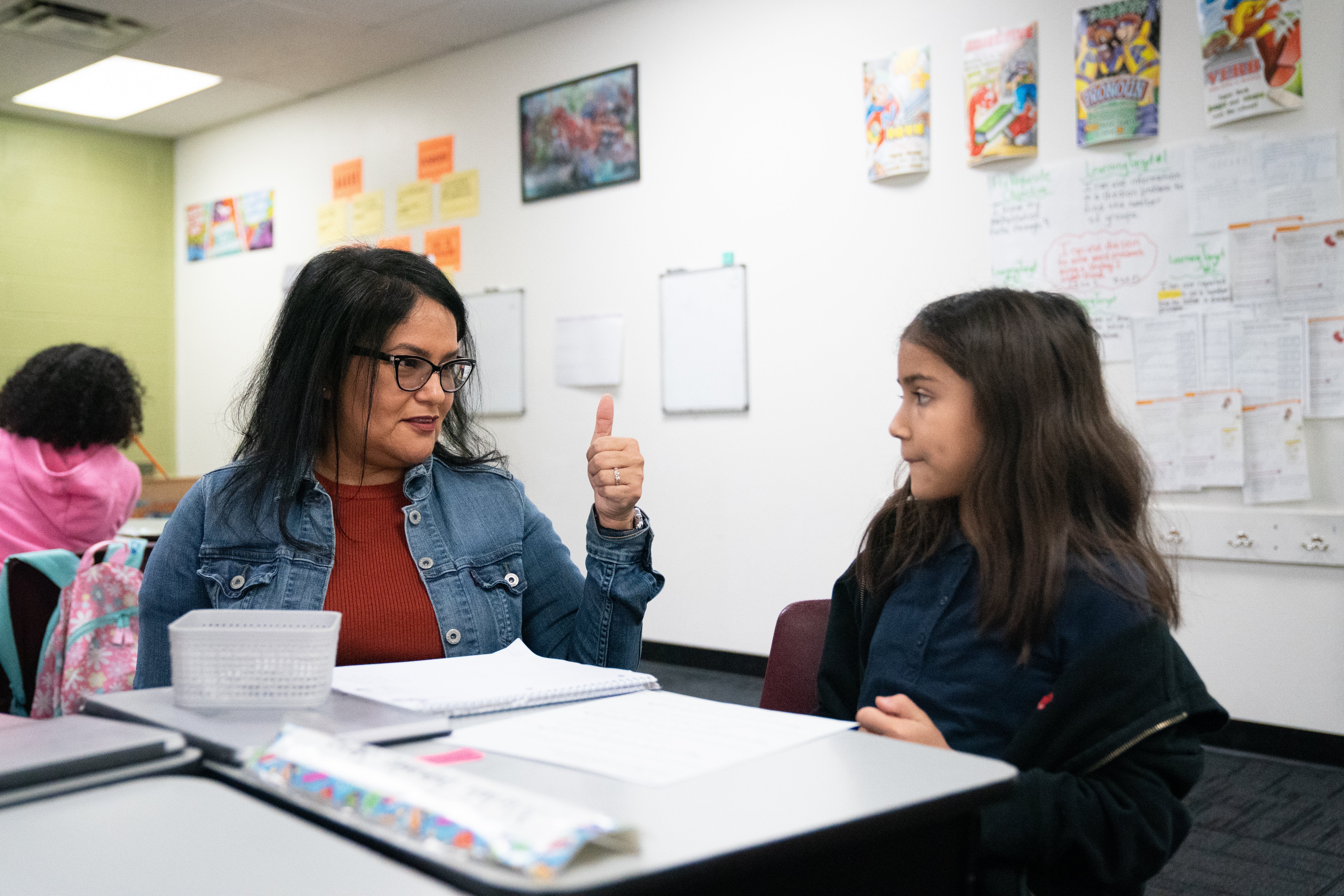 A teacher gives the thumbs up to a student as they sit together in a bright classroom